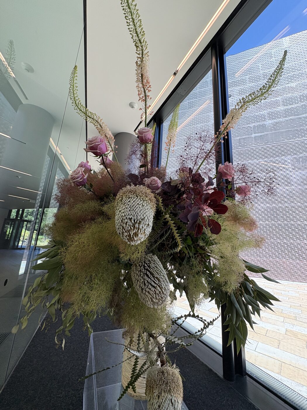 Floral arrangement of banksia, roses, and smoke bush in a sunlit atrium